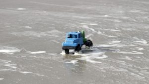 A blue toy truck navigates foamy ocean waves on Guaratuba Beach, Brazil.